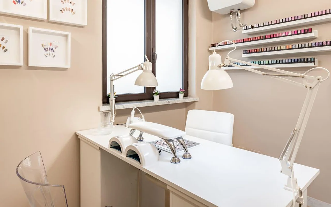 Nagų stotelė AG The Beauty Room's salone su švariu baltu stalu, dviem UV lempomis, reguliuojamomis darbo lempomis ir lentynomis, kuriose rikiuojasi eilės spalvotų nagų lakų. Ant šiltų atspalvių sienų kabo įrėminti nagų dizaino darbai.