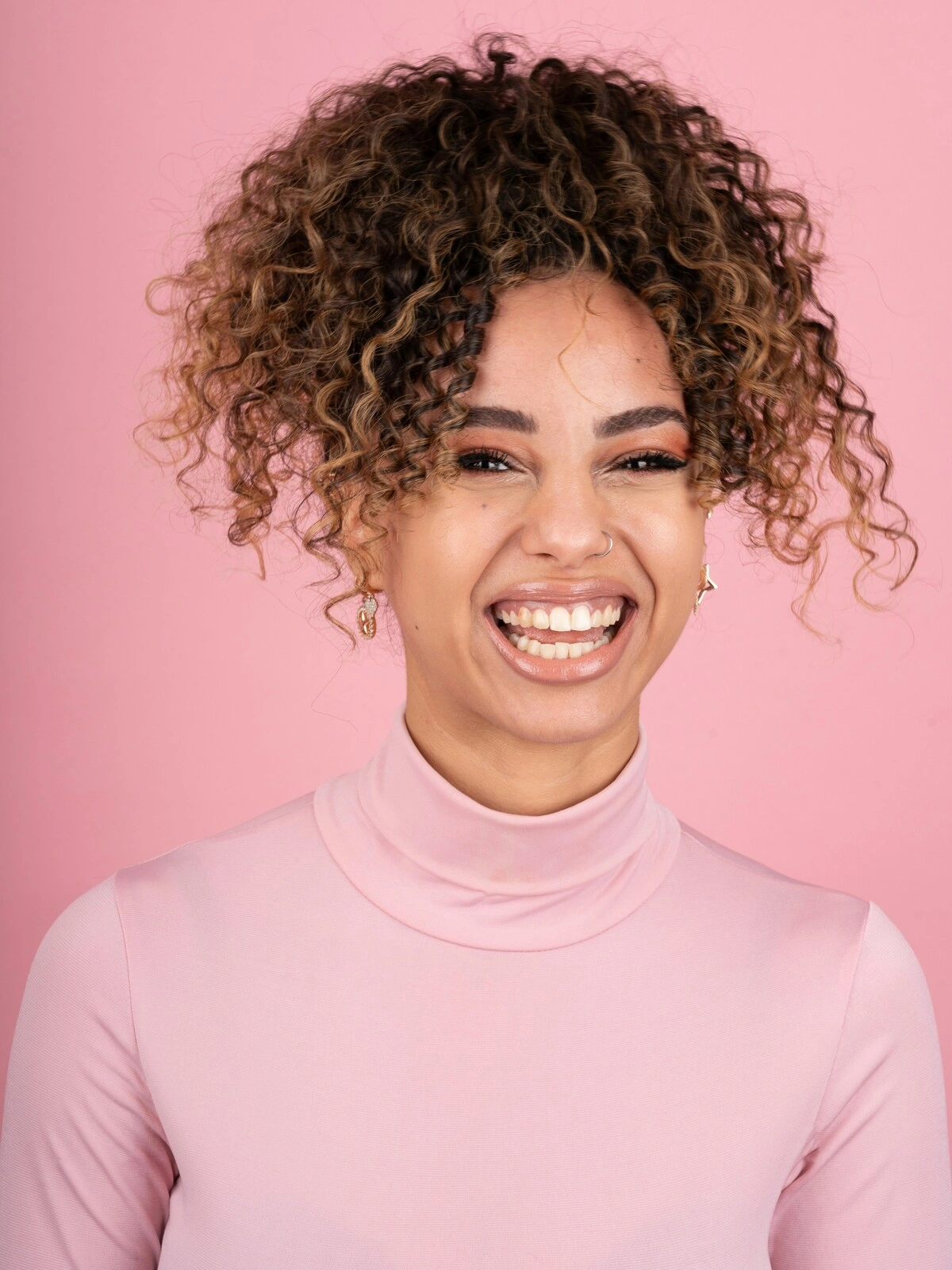 Studijinė nuotrauka besišypsančios moters su balayage ant afro plaukų, vilkinčios švelniai rožinį golfą
