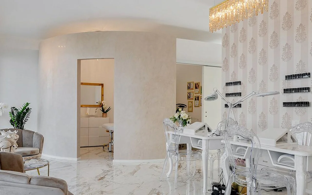 Elegantiškas MC Beauty Center interjeras su marmuro imitacijos grindimis, baltomis išlenktomis sienomis ir auksiniu krištolo sietynu. Dešinėje – nagų stotelė su skaidriomis kėdėmis, o pro auksu įrėmintas duris matyti procedūrų kabinetas su aksominiais foteliais.