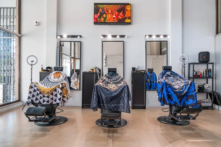 Šviesus aukštų lubų barbershopas su trimis juodomis kėdėmis, apdengtomis raštuotais apsiaustais, kiekviena prieš lemputėmis įrėmintą veidrodį, su žiediniais šviestuvais, sieniniu ekranu ir aukštais langais.