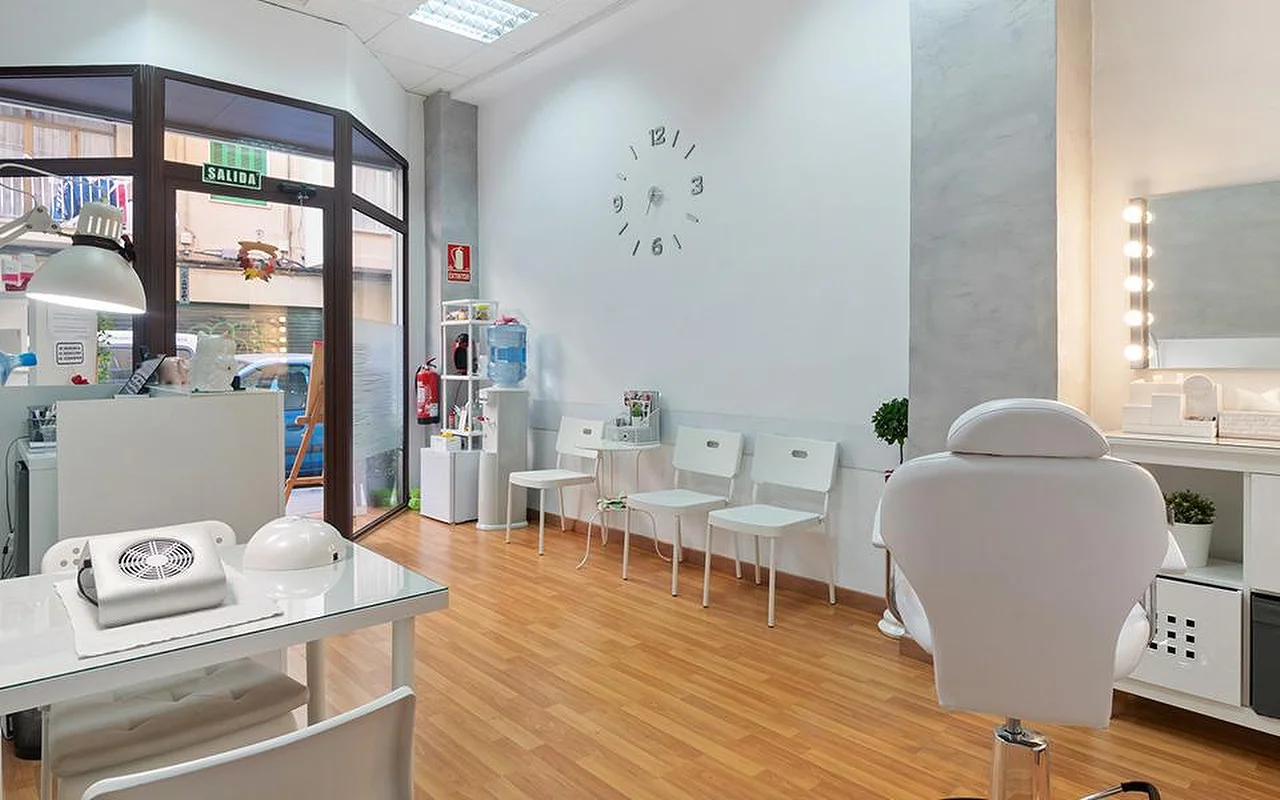 Šviesus baltas salonas su šviesaus medžio grindimis, balta laukiamojo kėdžių eile, minimalistiniu sieniniu laikrodžiu, apšviestu kosmetiniu veidrodžiu, baltu atlošiamu procedūrų krėslu ir manikiūro stalu prie lango.