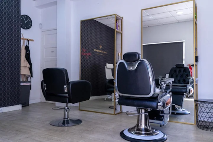 Santūrus salono interjeras su juoda kirpyklos kėde ir chromo bei odos barberio kėde ant šviesių medinių grindų, didelis auksinio rėmo veidrodis, sieninis laikrodis ir apsiaustai, kabantys prie tamsios akcentinės sienos.