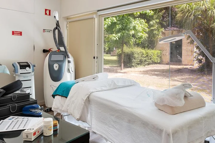 Procedūrų kambarys su balta paklota kušete prie nuo grindų iki lubų lango į žalią sodą, kūno procedūrų aparatu ant stovo, didinamąja lempa ir stikliniu šoniniu staliuku su priemonėmis.