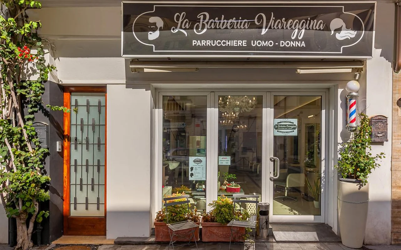 La Barberia Viareggina vitrina saulėtą dieną, su klasikiniu raudonai baltai mėlynu barberio stulpeliu stiklinių durų dešinėje. Įėjimą įrėmina vazonuose augantys augalai ir besikabinanti žaluma, o pro langus viduje matyti sietynas.