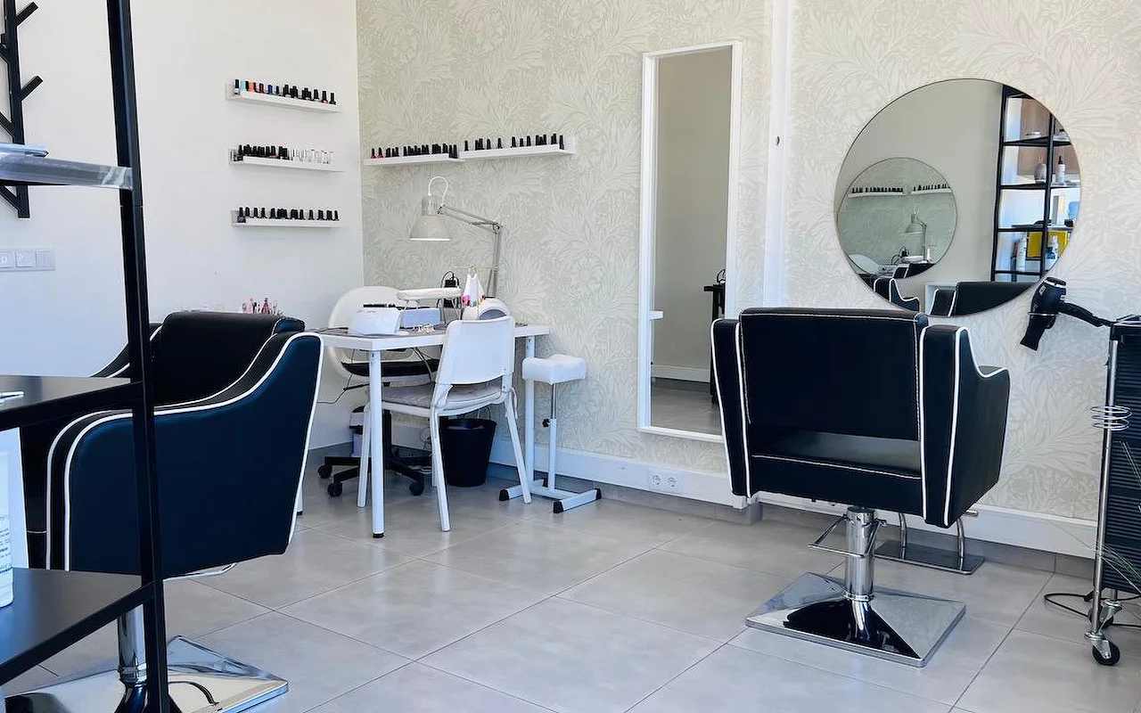 Kompaktiškas plaukų ir nagų salonas su juodomis formavimo kėdėmis, apvestomis baltai, apvaliu veidrodžiu ir aukštu veidrodžiu ant pilkų gėlėtų tapetų ir sieninėmis lentynomis, nusėtomis nagų lakais.
