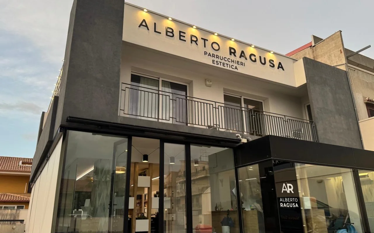 Alberto Ragusa išorė prieblandoje, dviaukštis salonas su didele apšviesta iškaba „Alberto Ragusa Parrucchieri Estetica“ virš nuo grindų iki lubų langų, su šilta šviesa, spindinčia iš vidaus.