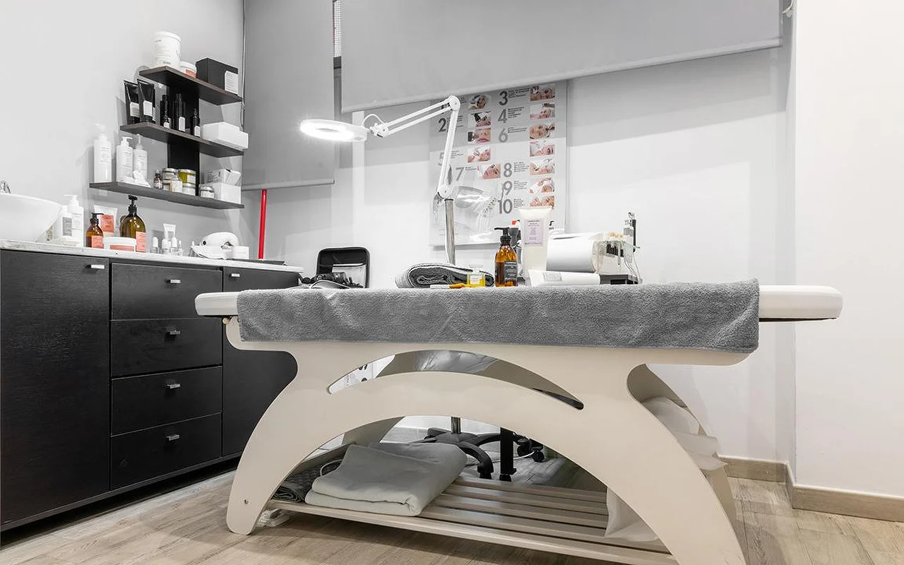 Kompaktiška kosmetologijos kabina Atelier Beauty and the Lab su kreminės spalvos gultu, reguliuojama lempa su didinamuoju stiklu, tamsiomis sieninėmis lentynomis, pilnomis odos priežiūros priemonių, ir sunumeruota procedūros žingsnių lentele ant sienos.