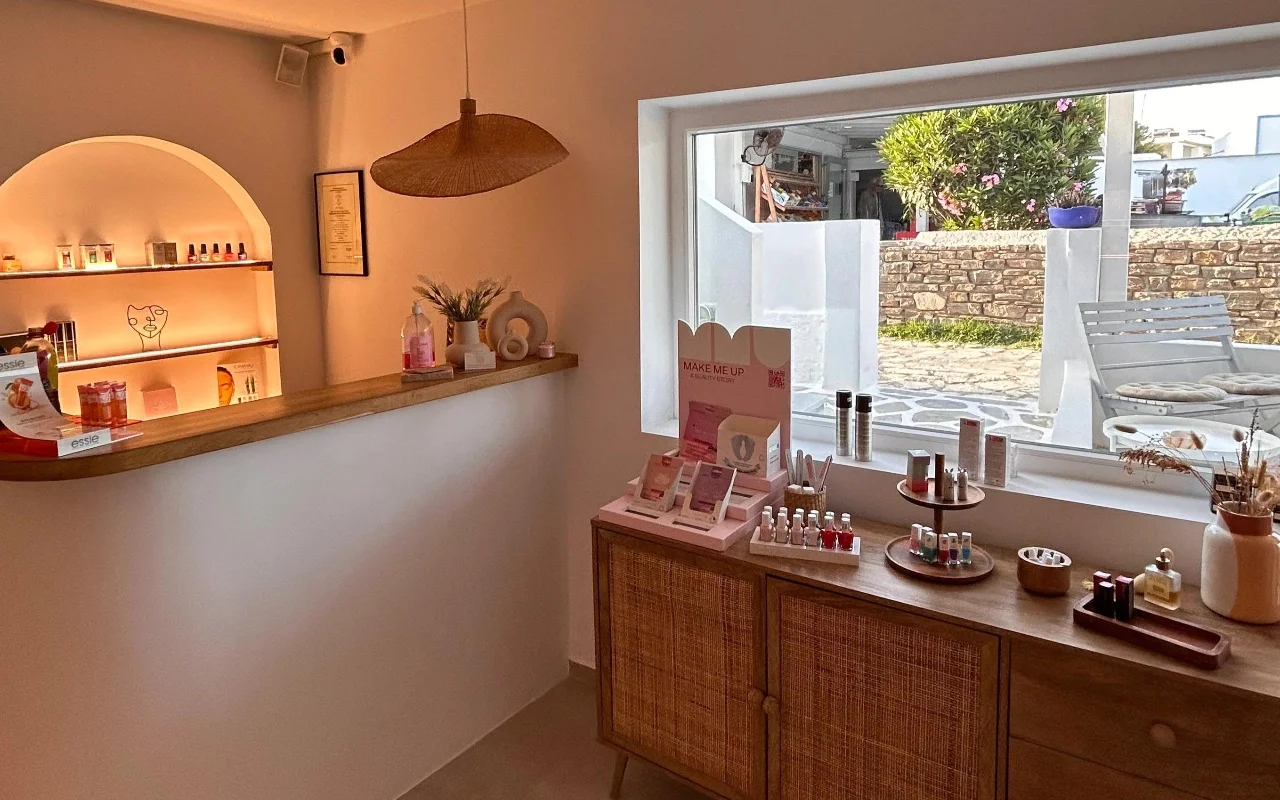 BeautiQ Esthetics and More interjeras Parose, su švelniai apšviesta arkine niša, pilna nagų lakų, pinta ratano pakabinamu šviestuvu ir dideliu langu, pro kurį matyti akmens siena ir saulėtas sodas.