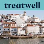 Baltai tinkuotas Algarve žvejų kaimelis su terakotinių stogų namais, kylančiais šlaitu virš ramaus vandens, mažos žvejų valtys, pririštos palei krantinę, palmės ir bažnyčios bokštas po giedru mėlynu dangumi.