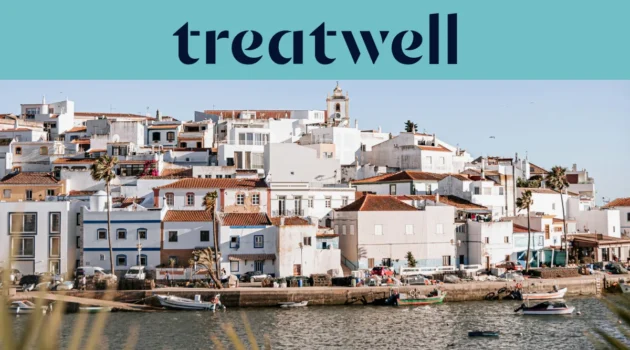 Baltai tinkuotas Algarve žvejų kaimelis su terakotinių stogų namais, kylančiais &scaron;laitu vir&scaron; ramaus vandens, mažos žvejų valtys, priri&scaron;tos palei krantinę, palmės ir bažnyčios bok&scaron;tas po giedru mėlynu dangumi.