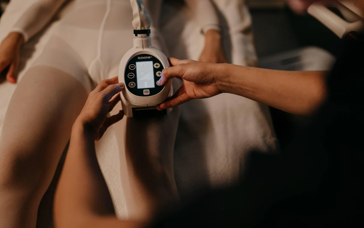 Kūno kontūravimo procedūra Beauty Room Romina Bongiovanni, kosmetologei laikant icoone aparatą prie kliento kojos su baltu procedūrų kostiumu, šiltoje, švelnioje šviesoje.