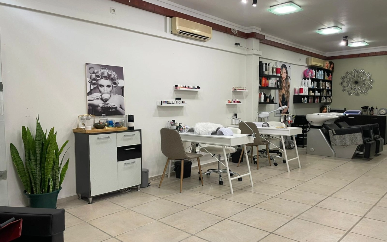 Plaukų ir grožio salonas su dviem baltais manikiūro staliukais ir smėlio spalvos kėdėmis, plovimo kriaukle ir produktų lentynomis palei sieną, aukšta sansevjera kampe ir didele nespalvota moters su plaukų suktukais nuotrauka.