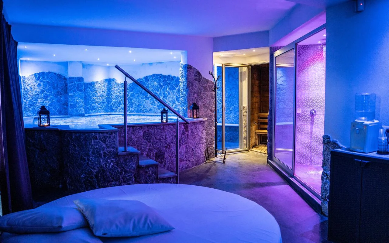 Privatūs spa apartamentai Bellezza e Benessere Taormina, apšviesti mėlynai ir violetiškai, su įleista sūkurine vonia akmens apdaila, medine sauna ir už jos mozaikinėmis plytelėmis išklota garine pirtimi, ir apvalia lova priekyje.