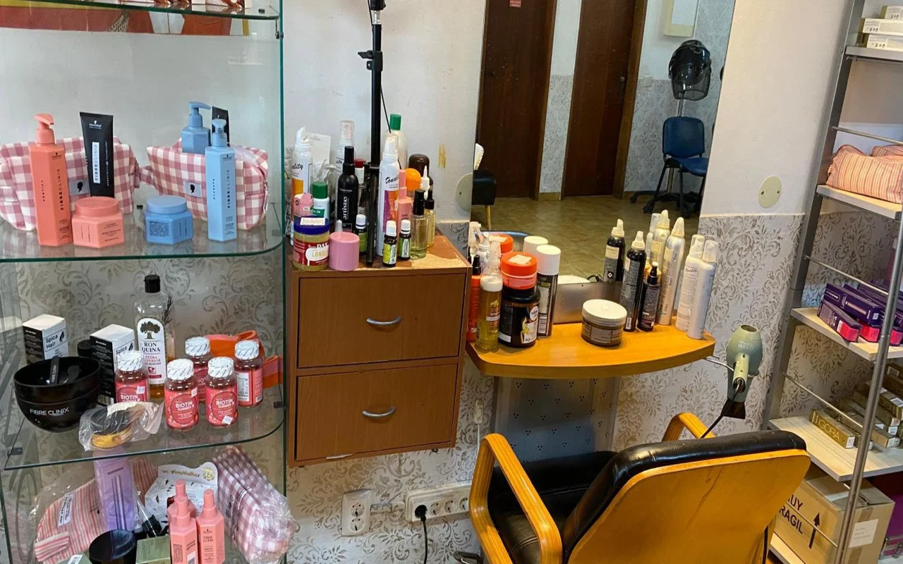 Kirpėjo darbo vieta su sieniniu veidrodžiu ir mediniu stalu, pilnu formavimo produktų, stikline vitrina su plaukų priežiūros produktais ir papildais šalia, ir klasikine medine bei juoda kirpyklos kėde.