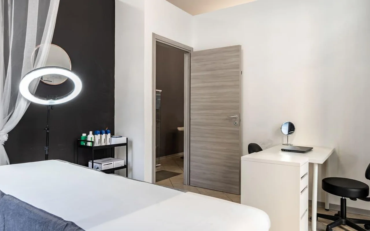 Procedūrų kabina Coquette Wellness and Beauty su balta lova, įjungta žiedine lempa ant stovo prieš juodą akcentinę sieną, lengvomis užuolaidomis prie lango ir mažu baltu stalu su kosmetiniu veidrodžiu prie durų, vedančių į atskirą vonios kambarį.