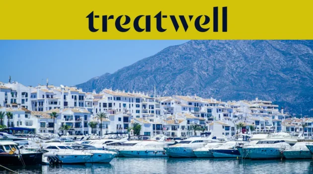 Puerto Banús prieplauka Marbella mieste, apsupta baltų daugiabučių ir pri&scaron;vartuotų jachtų su kalnais fone, po geltona Treatwell vėliava.