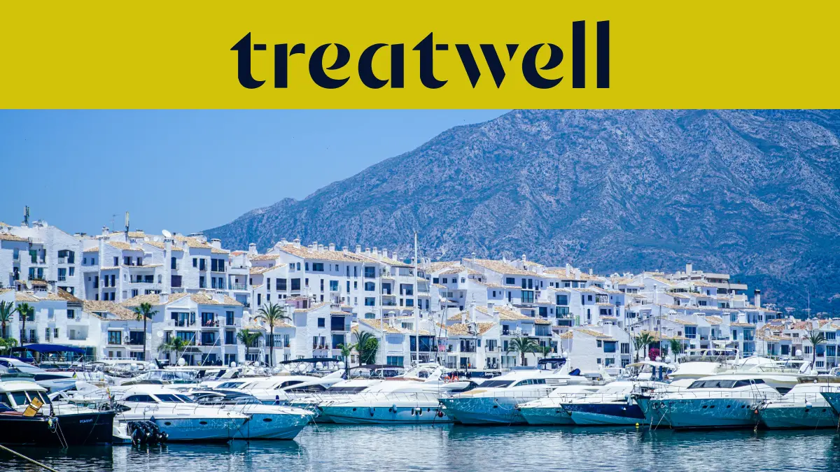 Puerto Banús prieplauka Marbella mieste, apsupta baltų daugiabučių ir prišvartuotų jachtų su kalnais fone, po geltona Treatwell vėliava.