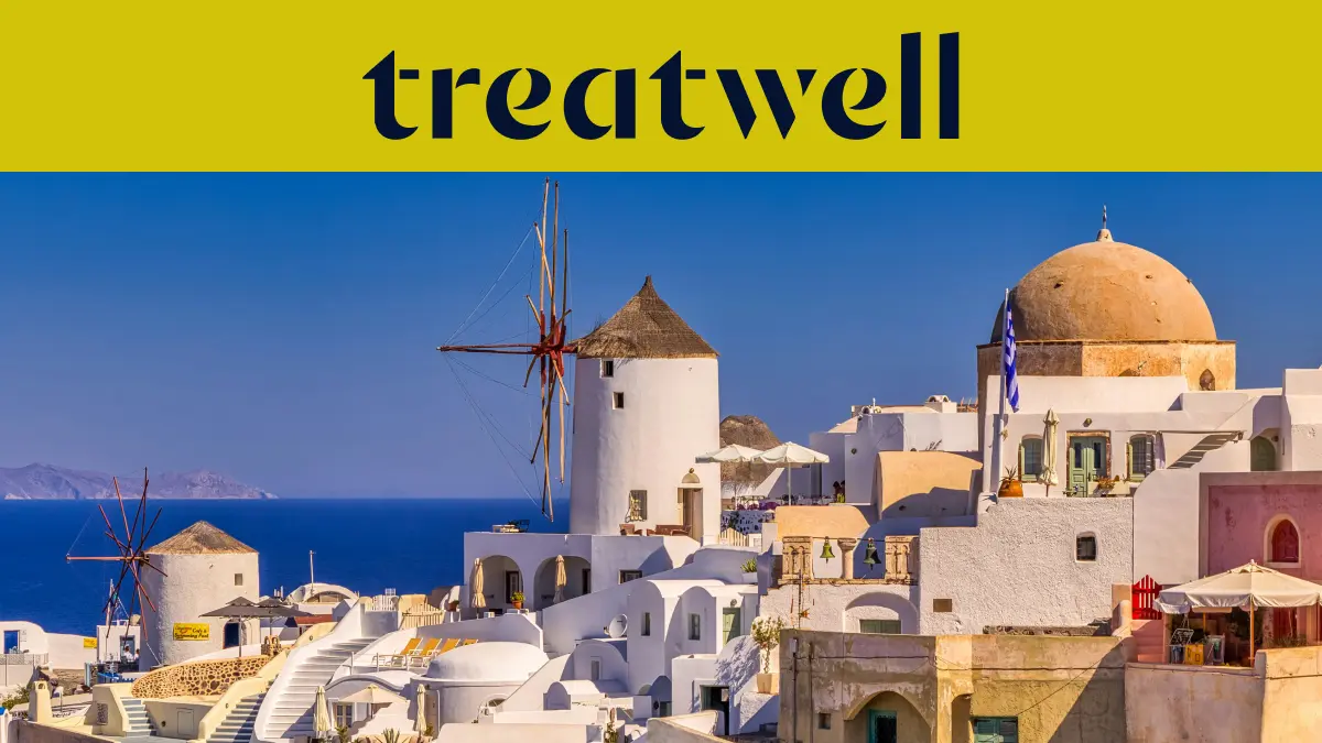 Baltai tinkuoti namai ir tradiciniai vėjo malūnai virš tamsiai mėlynos Egėjo jūros Oia miestelyje, Santorinyje, po geltona juosta su Treatwell logotipu.