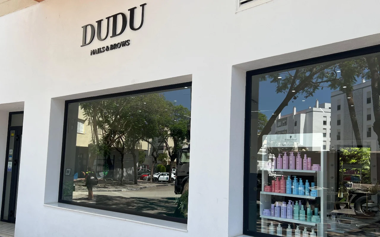 Baltas Dudu Nails and Brows fasadas Estepona mieste, pavadinimas tamsiomis raidėmis virš plačių vitrinų, kuriose matyti eilės plaukų priežiūros priemonių.