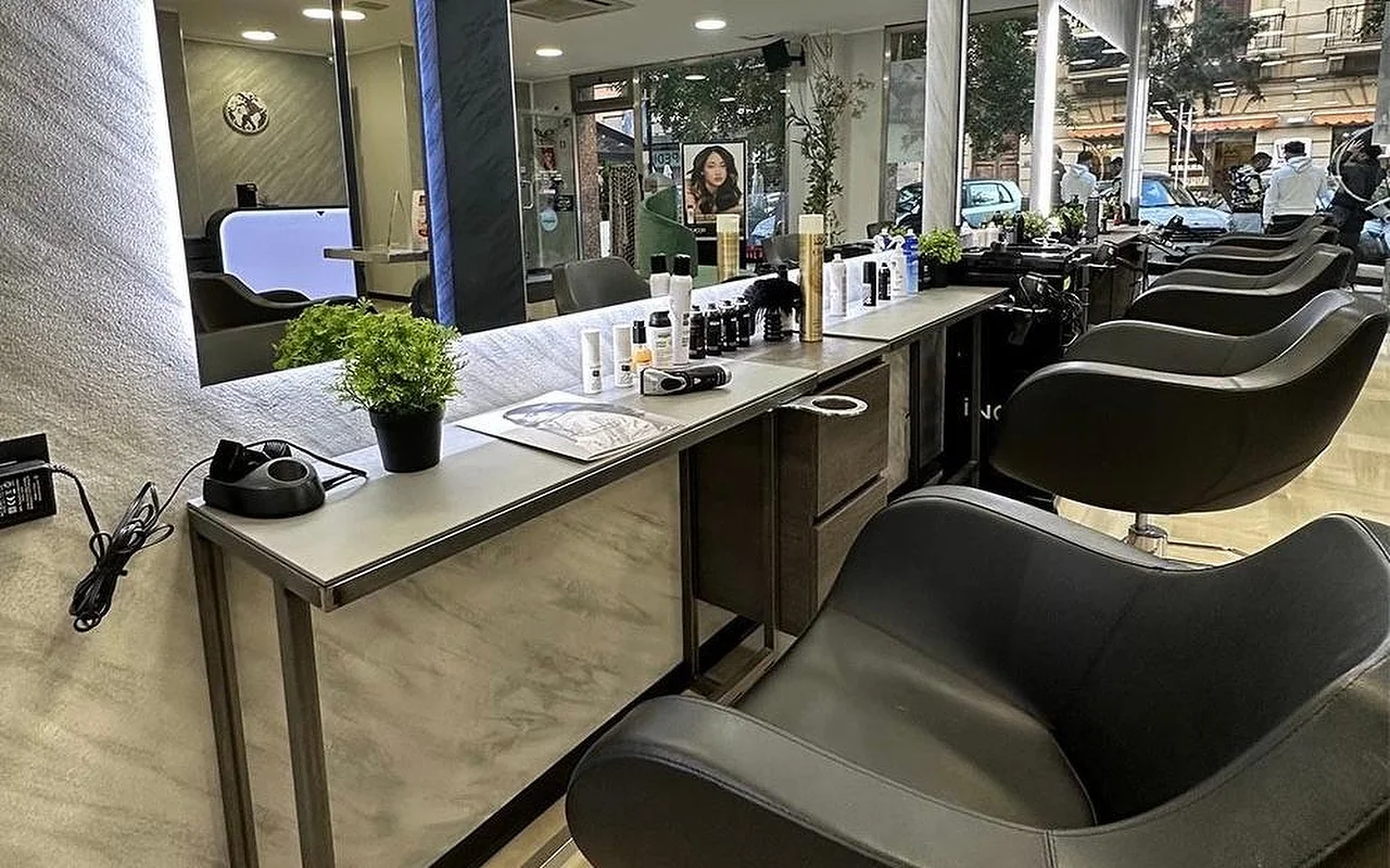 Equipe Beauty sušukavimo zona su juodų kėdžių eile palei marmuro imitacijos stalviršį, dideliu foniniu apšvietimu paryškintu veidrodžiu, formavimo priemonėmis ir vazonine gėle ant stalviršio, ir nuo grindų iki lubų langais į gatvę.
