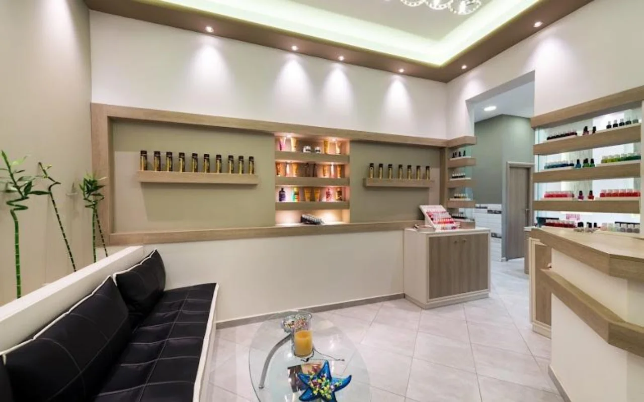 Kreminės spalvos Fabulous Nails and More registratūra Fira, Santorinyje, su juodu odiniu suoliuku, stikliniu staliuku, apšviestomis lentynomis su grožio produktais ir švelnia lubų šviesa.