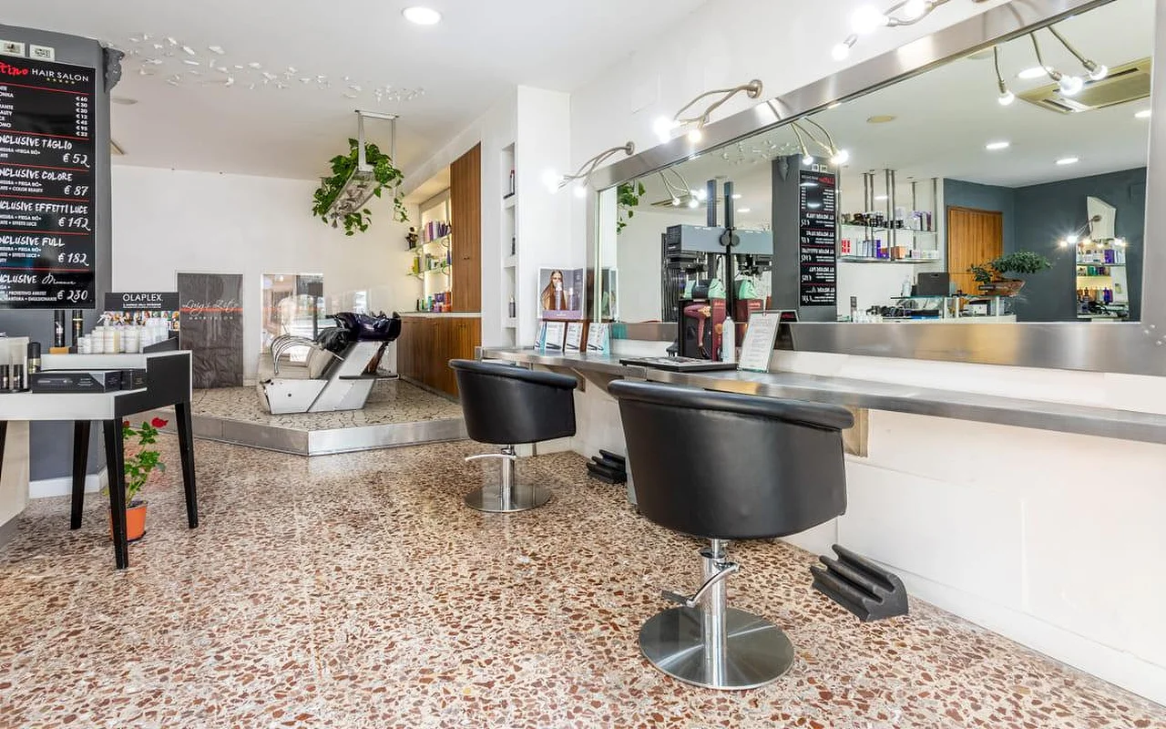 Hair Salon Luigi Zito vidus su terraco grindimis, dviem juodomis sušukavimo kėdėmis prieš ilgą veidrodį, kreidinėmis kainų lentelėmis ant sienos, produktų vitrina ir galvos plovykla gale.