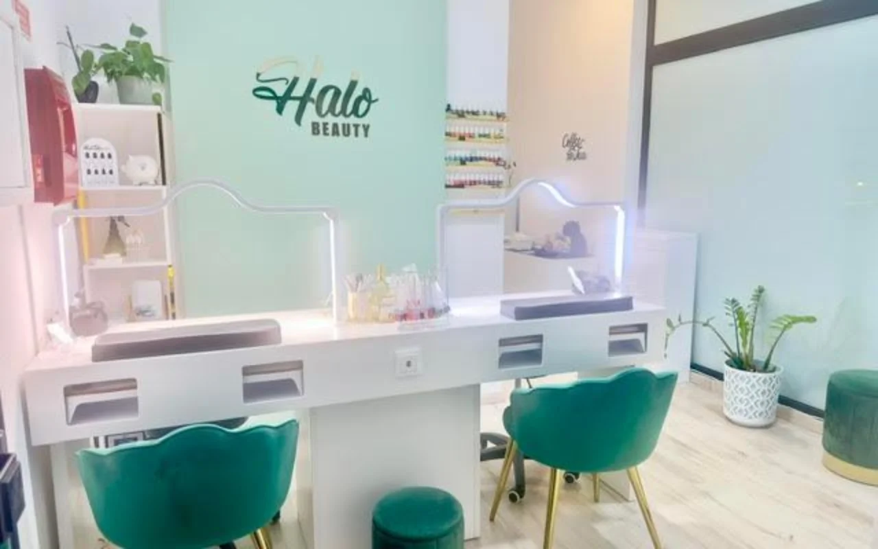Nagų studija su mėtinės spalvos akcentine siena ir Halo Beauty logotipu, dvigubu baltu manikiūro stalu su apšviestomis akrilo pertvaromis, dviem smaragdinės spalvos aksominiais foteliais, sieniniu nagų lakų stovu ir vazoninėmis gėlėmis.