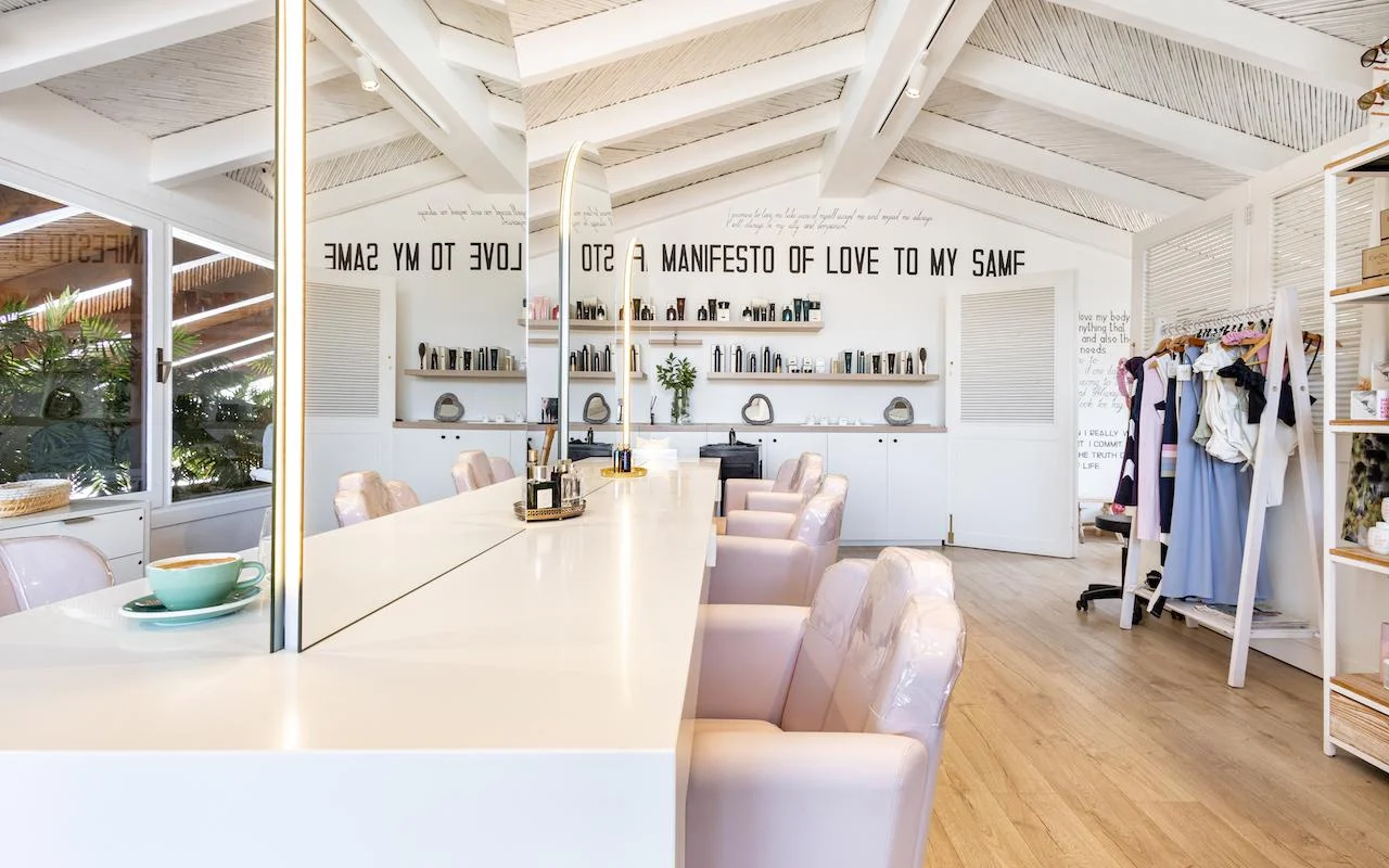 Šviesus Milk & Roses interjeras Puente Romano rajone, Marbella, su baltu formavimo pultu, šviesiai rožinėmis kėdėmis, sijų lubomis ir produktų lentynomis palei sieną.