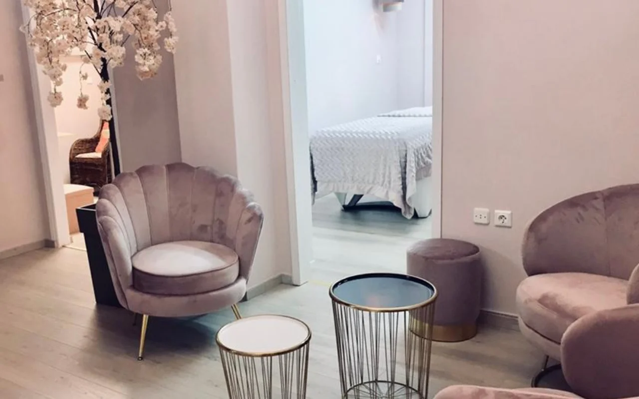 Pudros rožinės spalvos laukiamasis Nomikou Antonia Aesthetic Lab Sirose, su aksomine fotelio kėde banguotu atlošu, plonais auksiniais staliukais, žydinčia vyšnios šakele ir procedūrų kambariu už durų.