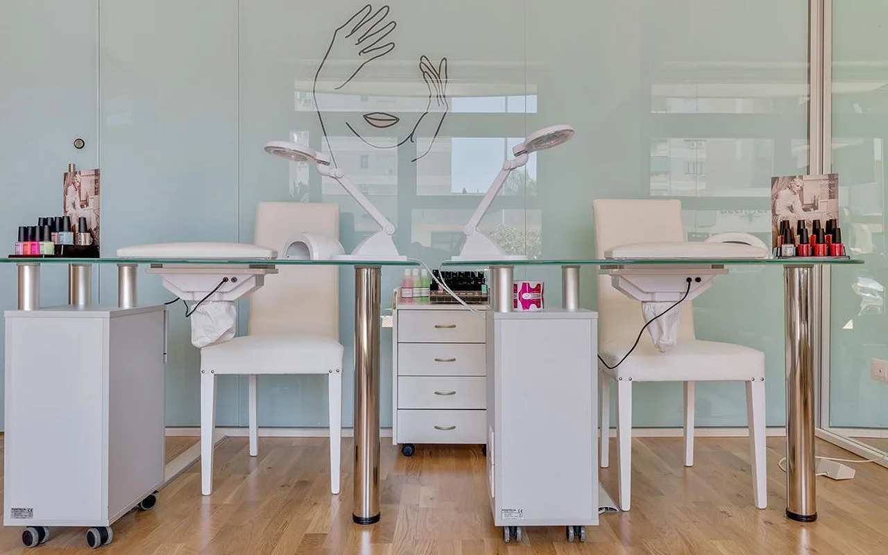 Nagų studija Perfetta Urban Beauty su dviem manikiūro staliukais stikliniu paviršiumi ir baltomis kėdėmis, darbo lempomis, spalvingomis lakų vitrinomis ir mėtų žalumo stikline siena su veido ir dviejų rankų linijine iliustracija.