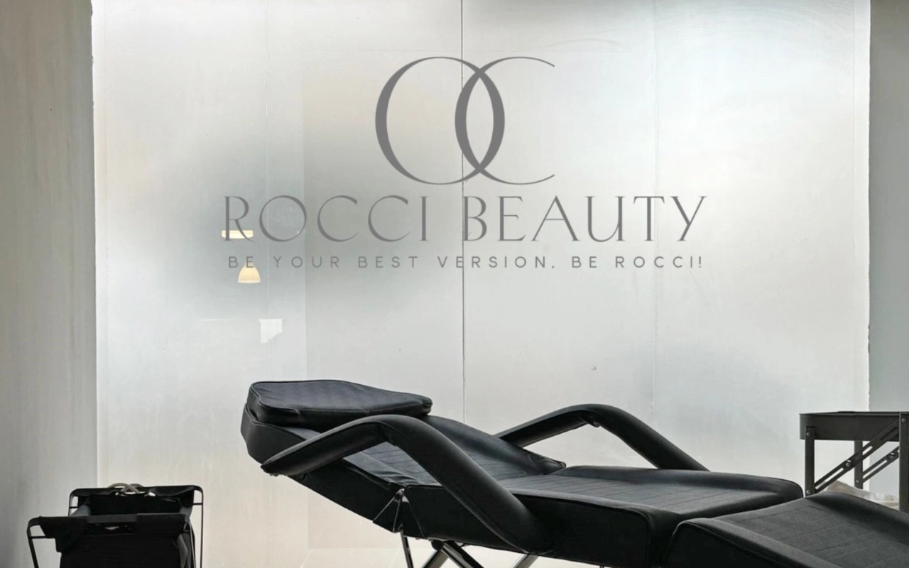 Procedūrų kambarys su juodu atlošiamu krėslu ir mažu įrangos vežimėliu priešais matinio stiklo sieną su Rocci Beauty logotipu ir šūkiu.