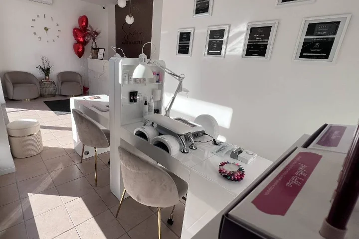 Baltas nagų salonas Sara Nails&Beauty Tortolì mieste su pilkomis aksominėmis kėdėmis ir įrėmintais sertifikatais ant sienos.