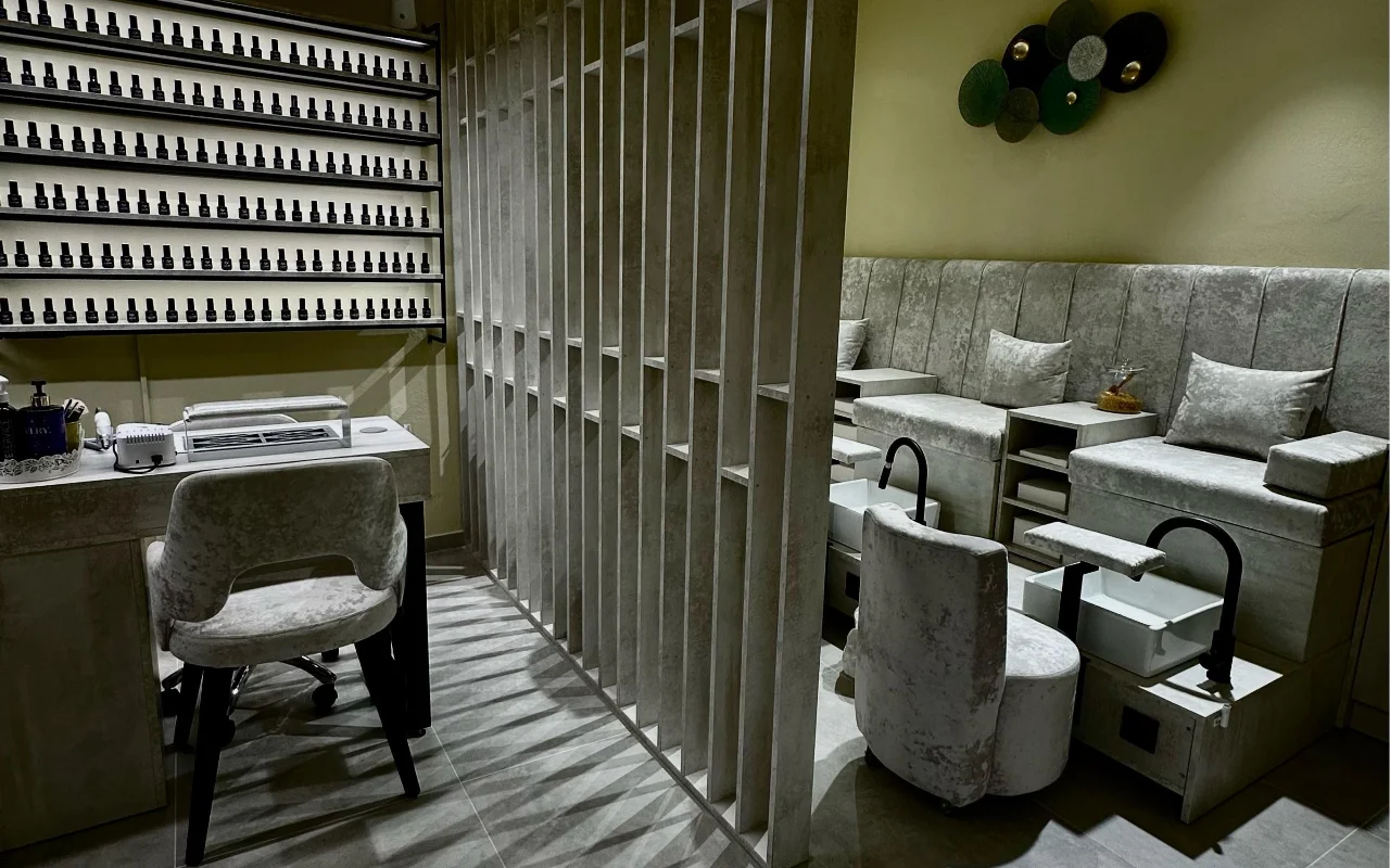 Procedūrų kambarys Sisu Nail Club and Academy Perissa, Santorinyje, su ištisa nagų lakų siena, pilkomis aksominėmis manikiūro ir pedikiūro kėdėmis ir ažūrine betonine pertvara.