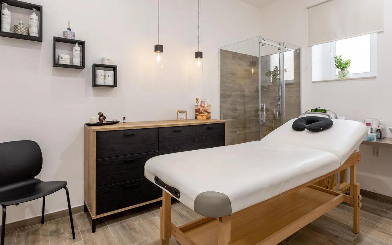 Masažo kambarys Thea Estetica su medine masažo lova su juodu veido atrama, komoda juodos ir medžio spalvos, dviem pakabinamais šviestuvais, kubinėmis sieninėmis lentynomis su produktais ir stikline dušo kabina su medžio imitacijos plytelėmis.