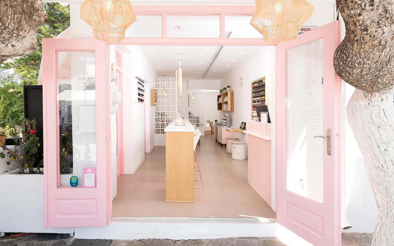 Rožine spalva įrėmintas Vivian Nail Boutique įėjimas Naoussa miestelyje, Parose, su atviromis durimis, pintais pakabinamais šviestuvais ir mediniu prekystaliu, baltai tinkuotame kikladiško stiliaus pastate šalia medžio.