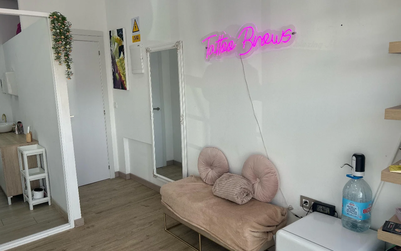 Procedūrų erdvė YG Beauty Estudio salone Torremolinos mieste, su rožine „Tattoo Brows“ neonine iškaba, viso ūgio veidrodžiu ir minkšta smėlio spalvos kušete.