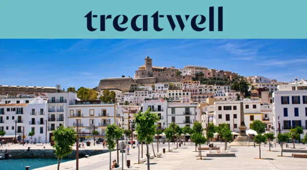 Saulėta panorama į Ibiza miestą, kur baltai i&scaron;balinti pastatai kyla iki katedros ir senųjų Dalt Vila įtvirtinimo sienų ant kalvos. Medžiais apsodinta uosto promenada eina pirmame plane po skaidriai mėlynu dangumi.